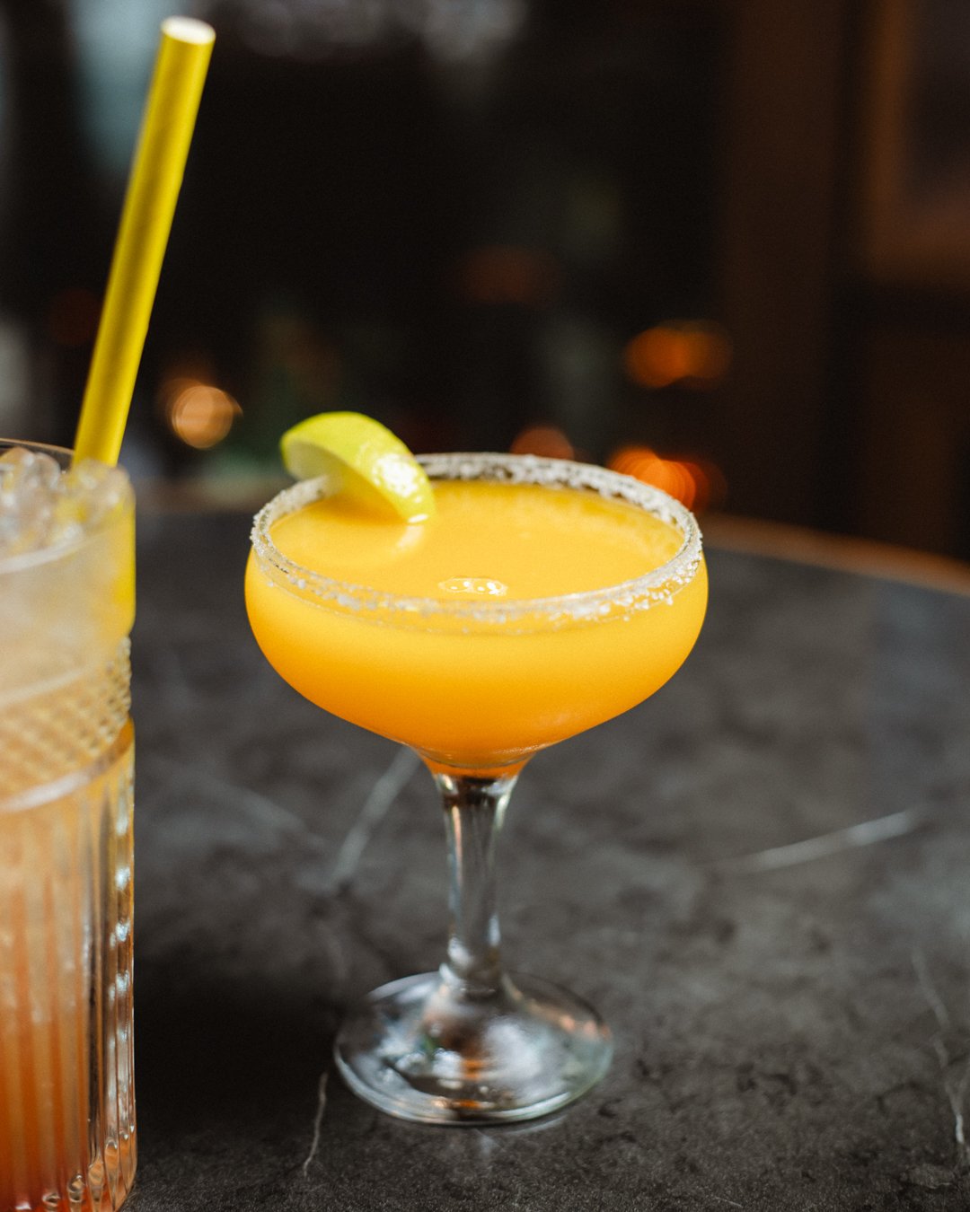 A frozen mango-gold cocktail in a salt-rimmed coupe with a lime wheel, warm bar lights behind