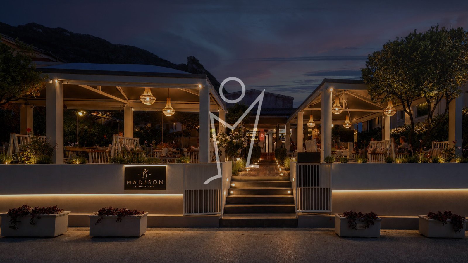 Madison Garden at dusk, terrace lit with warm lanterns beneath the Agios Gordios hillside